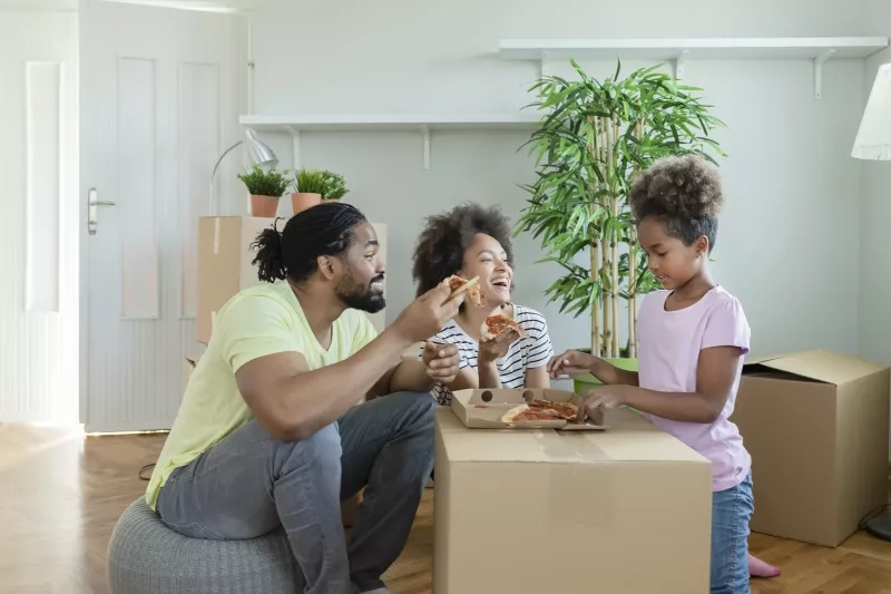 Young happy family have a pizza lunch break on the floor after moving into a new home with boxes around them