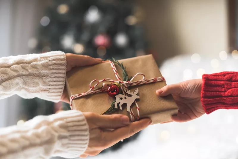 Woman in sweater giving a wrapped Christmas gift box to child. Glowing snow bokeh, fir tree. Winter holidays.