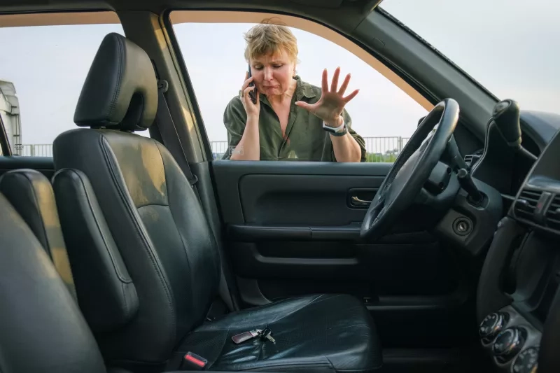 woman looks through the glass into the interior of her car with the keys in the driver's seat. 