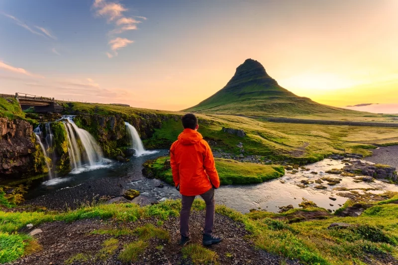 Majestic landscape of Godafoss waterfall flowing with colorful sunset sky and male tourist standing at the cliff on Skjalfandafljot river in summer at Northern Iceland