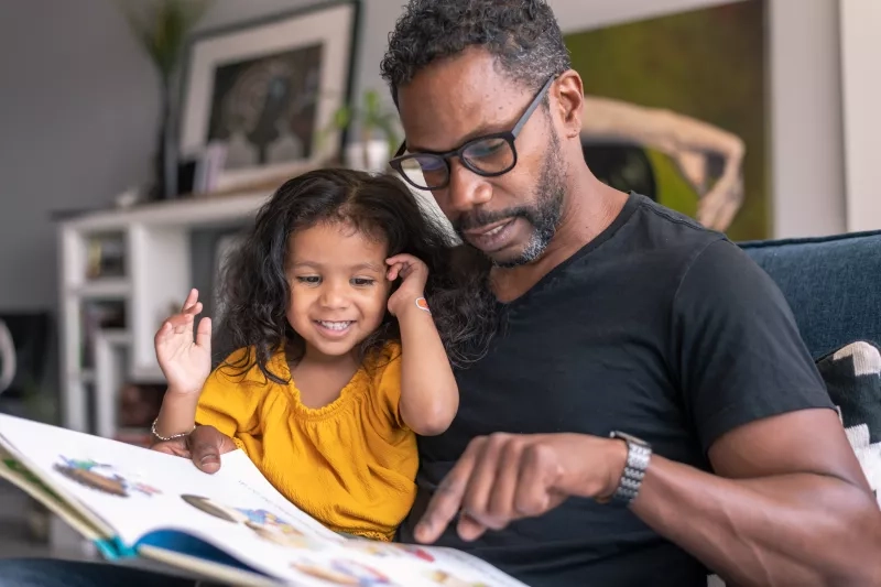 Child sitting on her father's lap and is smiling while looking at the book.