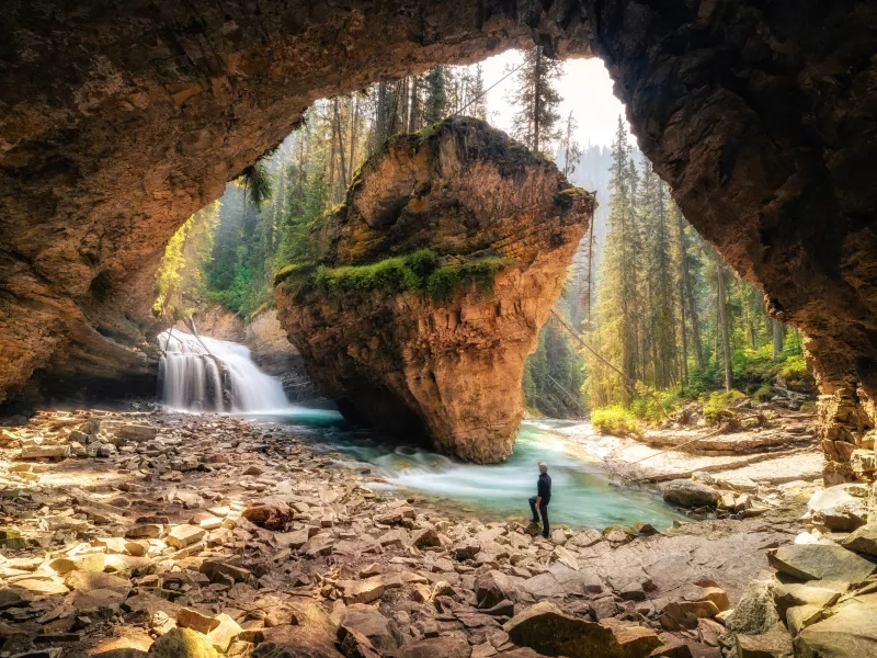 Man Standing In Cave Against Waterfall At Forest
