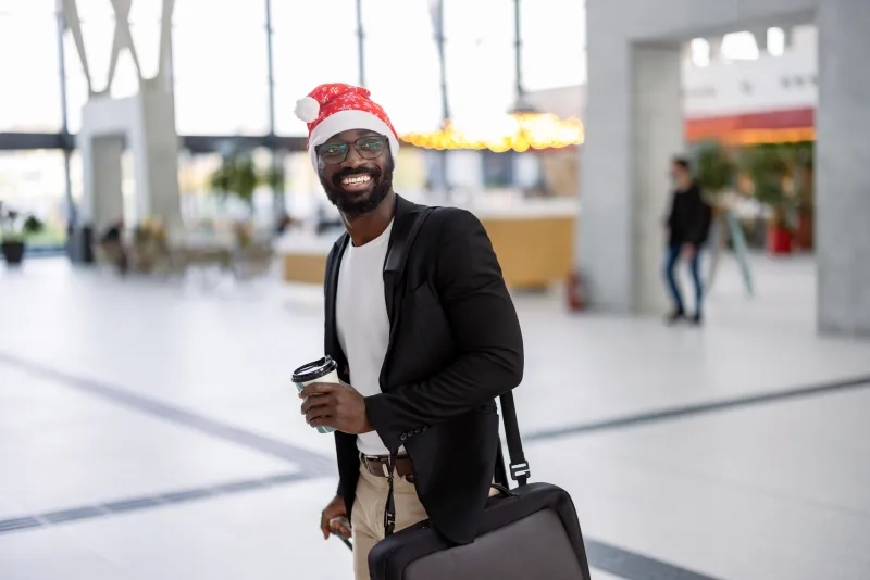 A joyful man sporting a Santa hat carrying a coffee and pushing his suitcase