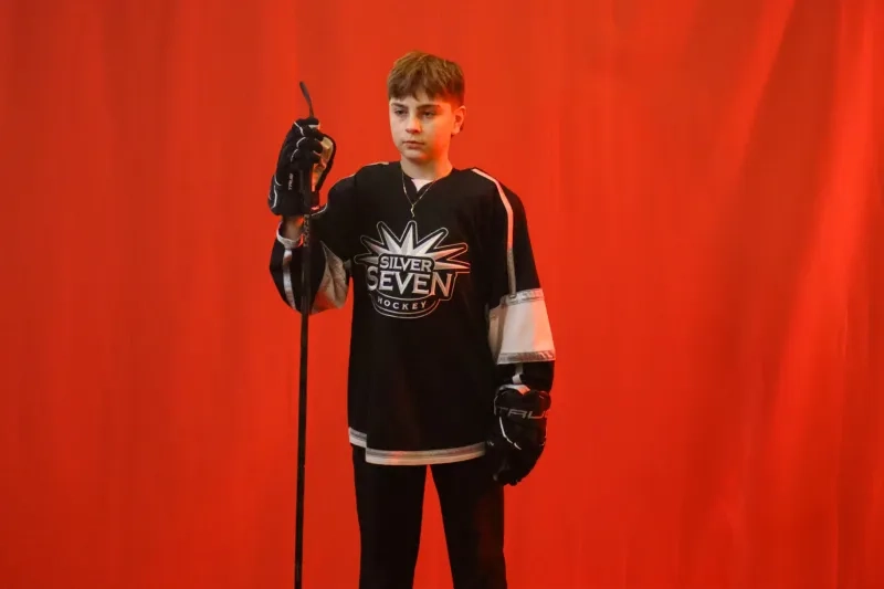 A young teen boy poses before a red backgroun in hockey jersey carrying his hockey stick