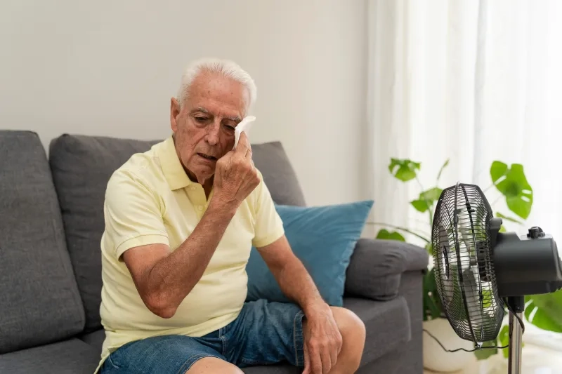 Senior man wiping sweat from face while sitting on sofa near an electric fan