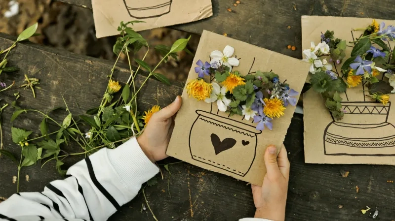 A child's hands are holding a piece of cardboard with a drawing of a vase filled with real wildflowers and leaves