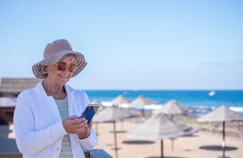 senior woman with hat and sunglasses sitting relaxed near the beach using her smartphone