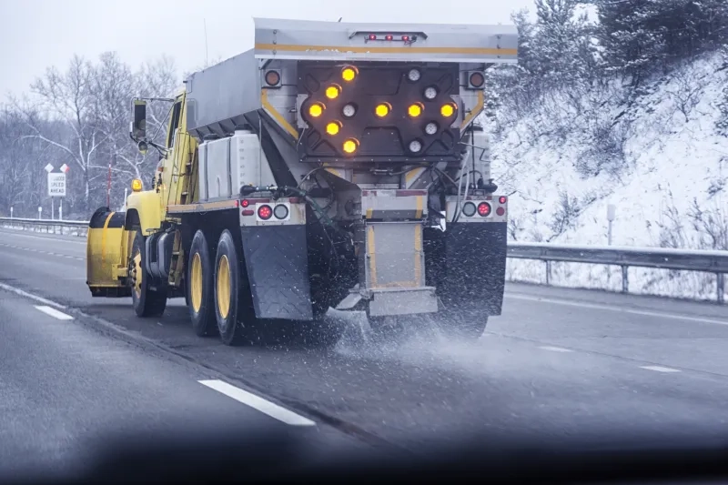 Express passing lane car driver POV about to move past a state transportation department snowplow dump truck driving in the center lane on the New York State Thruway highway. The large commercial vehicle has a bright, flashing emergency light directional arrow pointing to the left attached on the back of the rig to warn faster following traffic to pass on the left while it spreads and scatters road salt with its automatic spreader equipment. Early February winter weather in western New York State, USA.