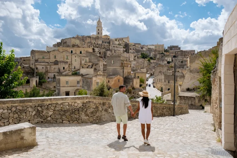 A couple strolls hand in hand along a cobblestone path, surrounded by the ancient cave dwellings of Matera, Puglia. The sun illuminates the historic architecture beneath a sky filled with clouds.