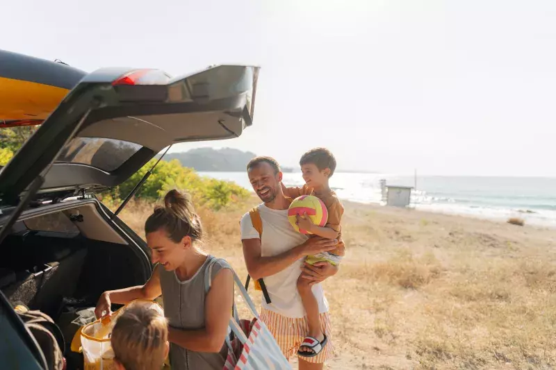 A family is at the beach with their car's trunk open. A man in a white tank top is holding a child, both smiling. A woman is arranging items in the trunk while another child stands nearby. The beach and ocean are visible in the background under a clear sky.