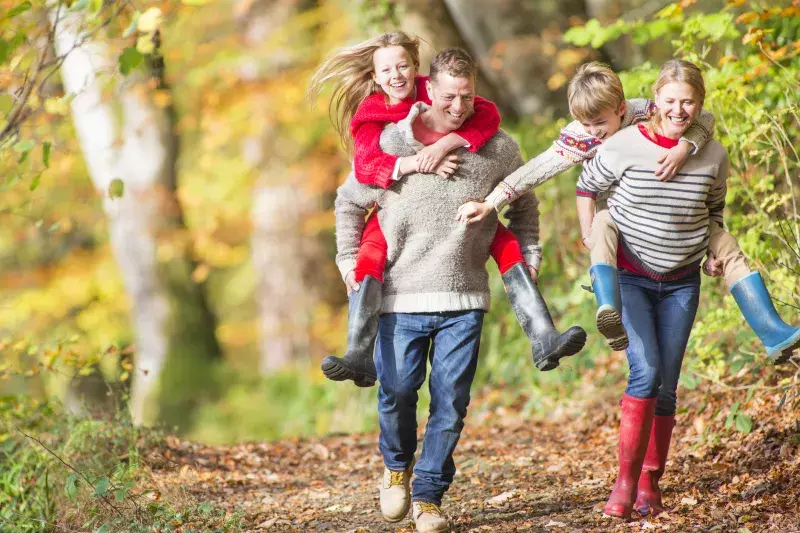 A happy family mess about in the woods together. The mother and faher give their children piggybacks.