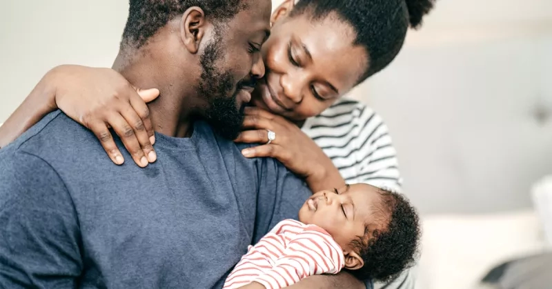 A loving family moment captured as a father holds a sleeping baby in his arms. The mother leans in, embracing both with a tender smile. The baby is dressed in a red and white striped outfit.