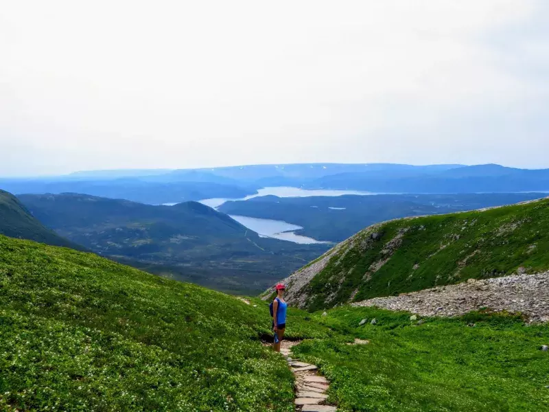 A young female hiker climbing near the summit of Gros Morne Mountain, in Gros Morne National Park, Newfoundland and Labrador, Canada.  A gorgeous green valley of mountains and lakes are behind her.