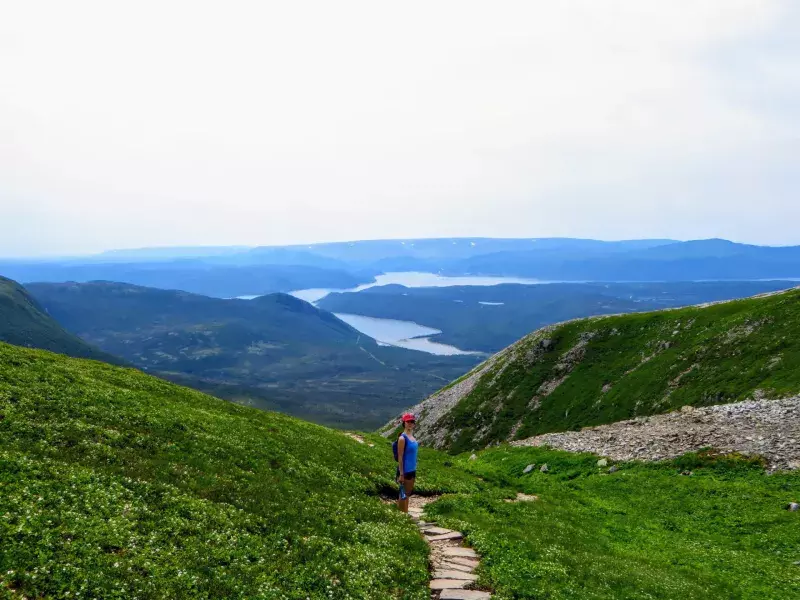 female hiker climbing near the summit of Gros Morne Mountain, in Gros Morne National Park, Newfoundland and Labrador, Canada.  A gorgeous green valley of mountains and lakes are behind her.