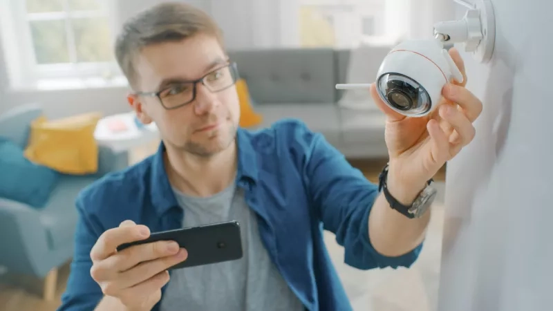 Man in Glasses Wearing a Blue Shirt is Adjusting a Modern Wi-Fi Surveillance Camera with Two Antennas on a White Wall at Home. He's Checking the Video Feed on his Smartphone.