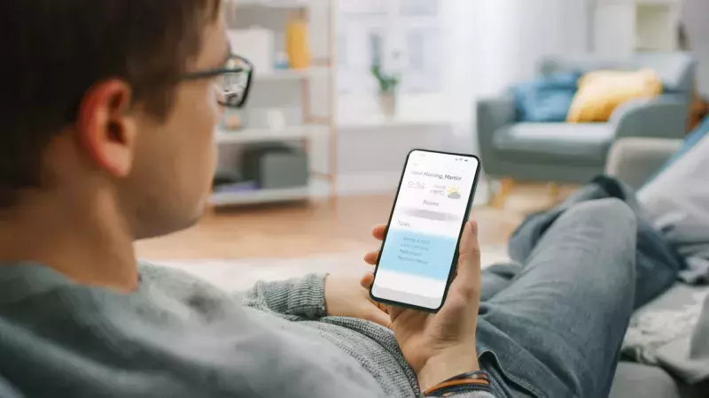 Over the Shoulder Shot of a Young Man at Home Sitting on a Sofa and Using Smartphone for Checking Weather Predictions and Coming Day Plans in Daily Schedule Calendar. Cozy Living Room.