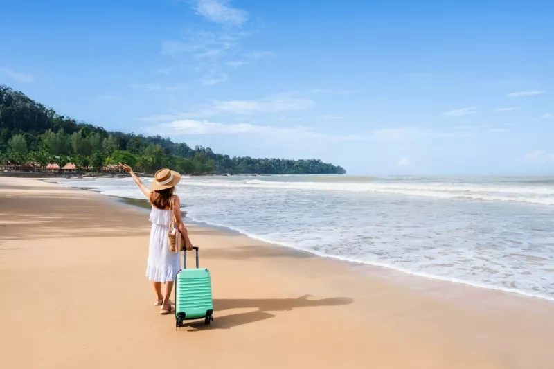 Young woman traveler with luggage relaxing and enjoying on tropical beach in sunny day, Summer vacation and travel concept