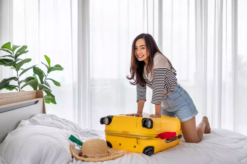 Woman packing and trying to close luggage, smiling. Girl preparing suitcase for summer vacation travel trip, looking at camera.