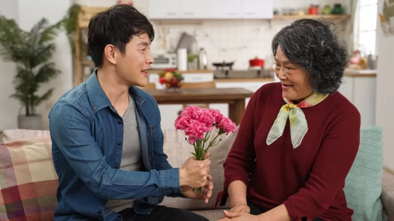 cheerful asian adult son surprising mother with carnation bouquet on Mother's Day. happy senior woman embracing the man with smile on sofa at home