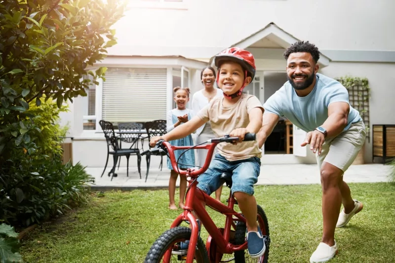 Learning, bicycle and proud dad teaching his young son to ride while wearing a helmet for safety in their family home garden. Active father helping and supporting his child while cycling outside